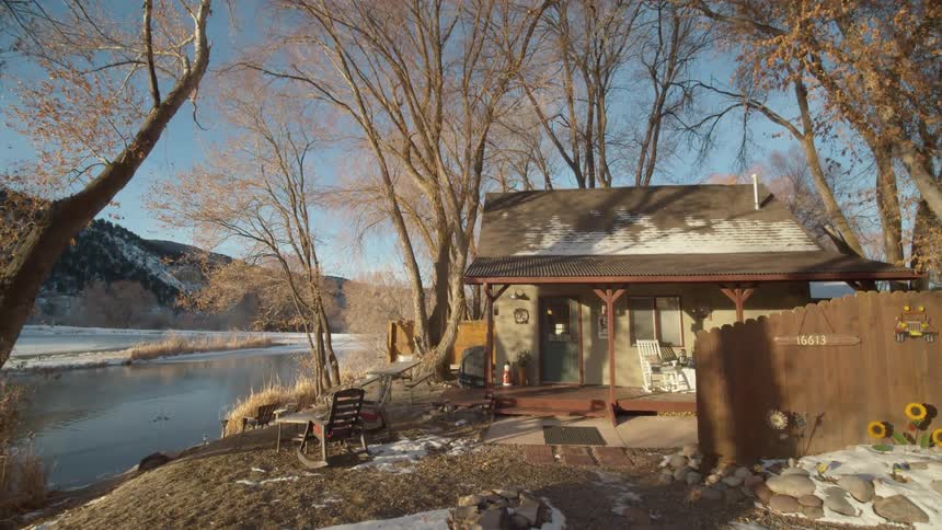 A cabin by a river during the winter with mountains in the distance