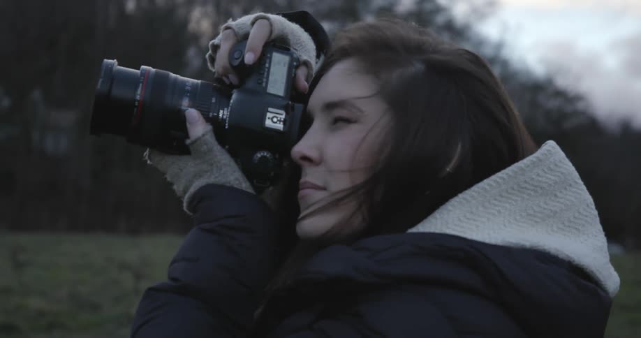 A young woman standing in a field in warm clothes taking photos with a forest in the background
