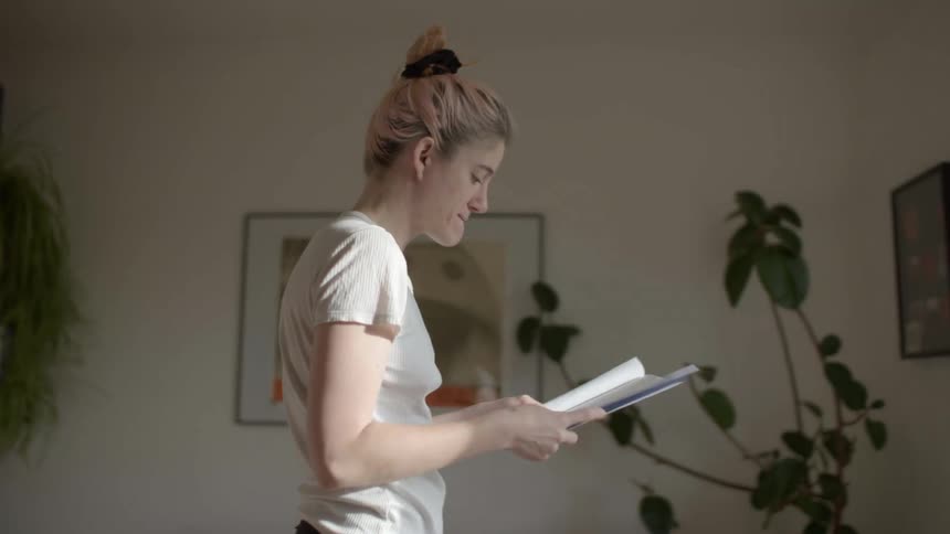 A woman paces in a room while reading a booklet in her hands during the day