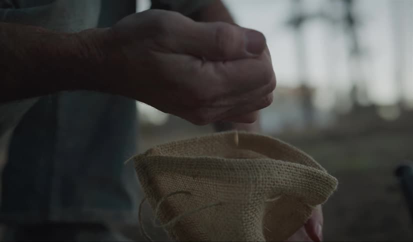 A man kneeling outdoors during the day takes a handful of seeds from a burlap sack before letting them fall back into the sack
