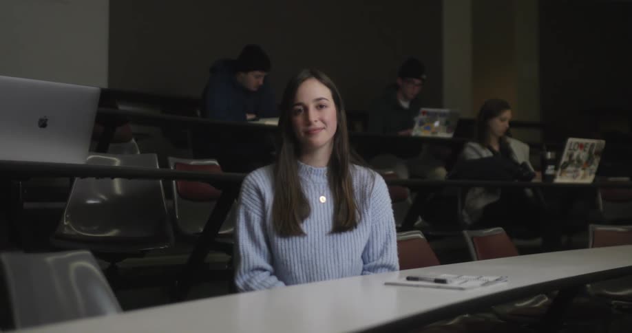 A woman sitting at an elongated table in a study hall smiles at the camera