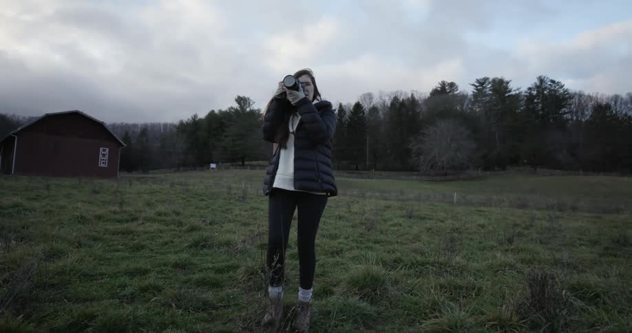 A young woman walking in a field with a forest behind her taking picture with a camera