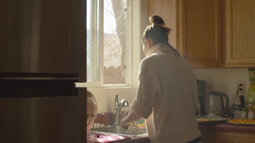 Woman wearing sweater washes dishes near window showing leafless tree