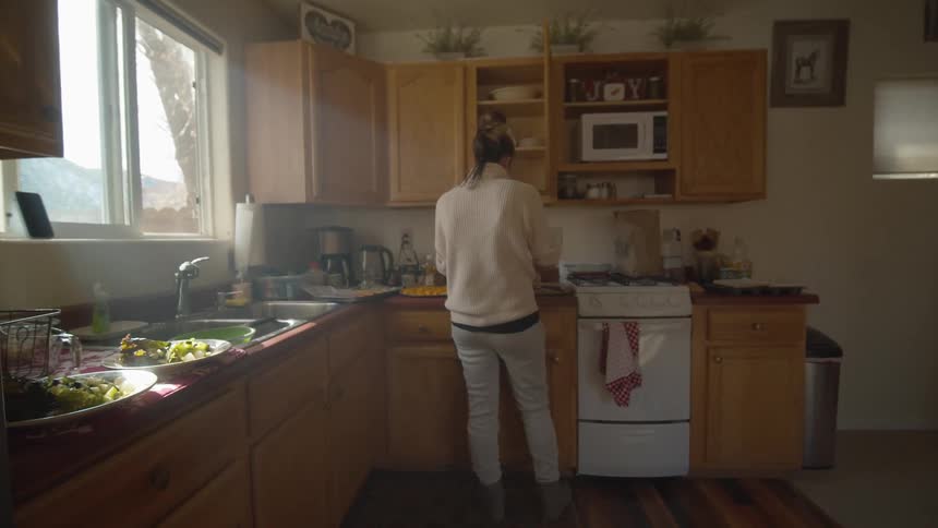 Woman prepares food in kitchen as sun shines through window showing leafless tree