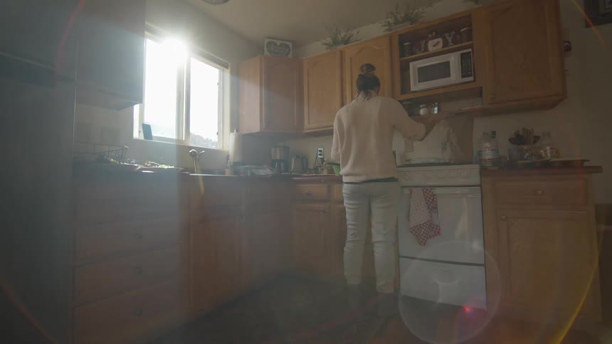 A flare of sunlight beams into a small kitchen window where a woman is preparing food on a countertop