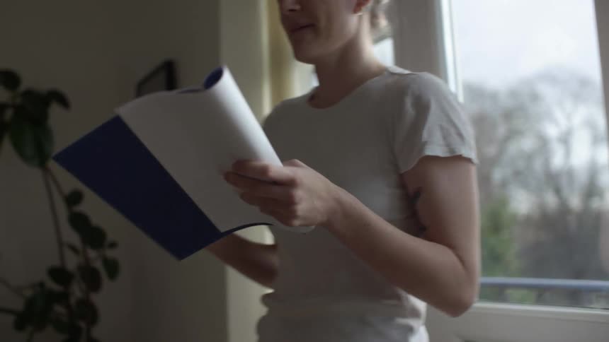 A woman in a room pacing and talking with a book and a pen in her hands