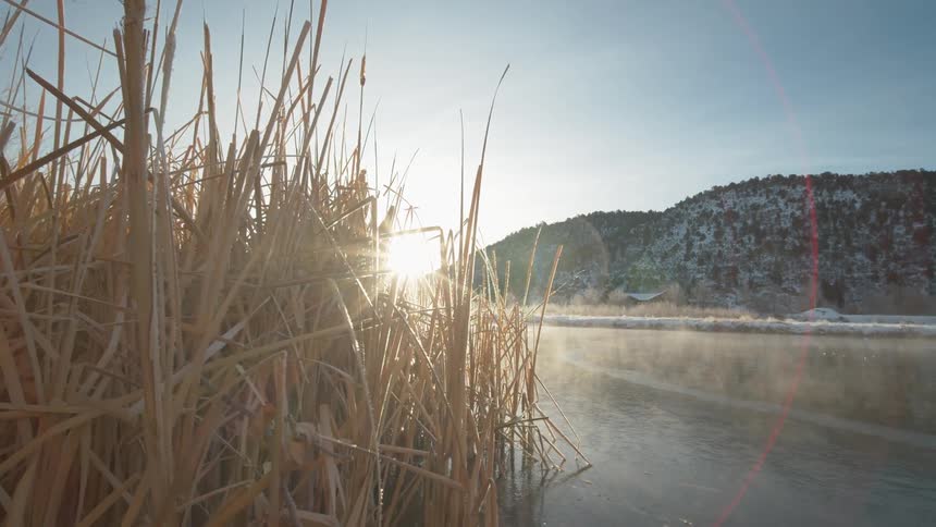 An outdoors scene featuring the sun rising a river a mountain with light snow on it and dried grass along the shore of the water