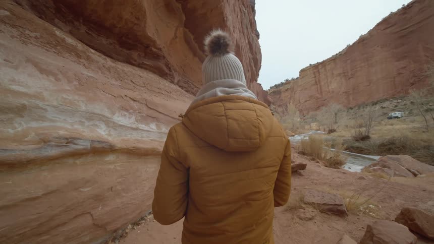 A person with a jacket and a hat walking in a canyon with the sky and clouds in the background