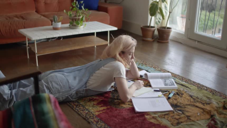 A teenage girl lies on the floor at home and does homework