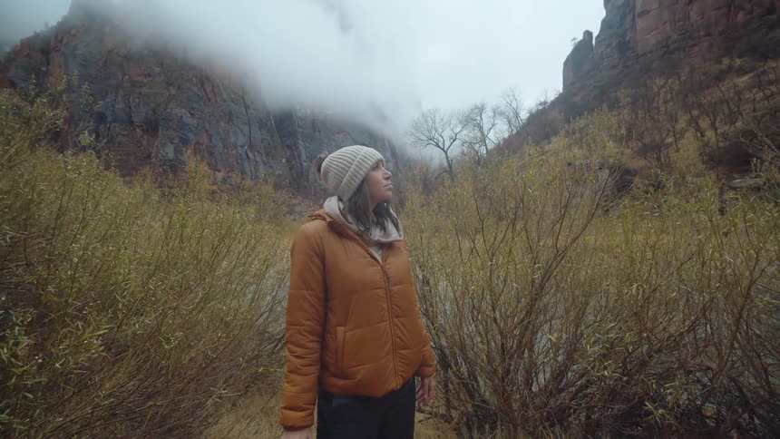 A woman with a hat and coat holding a camera with trees a canyon the sky and clouds in the background