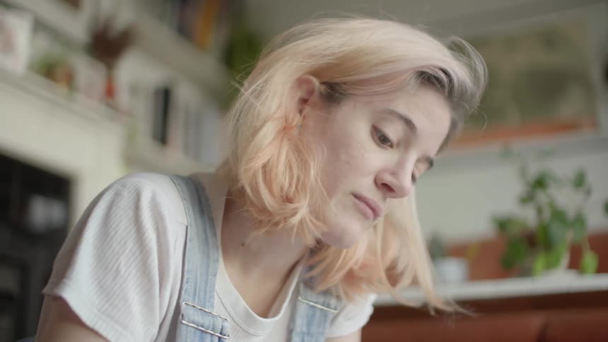 A blonde woman in a white tee shirt and overalls writing indoors