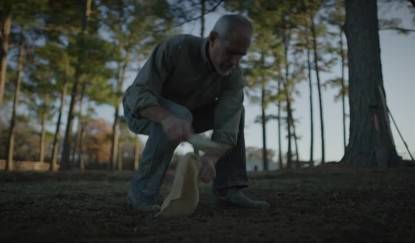 A man walks to a clearing amidst some trees and crouches down to begin digging a hole with a hand trowel