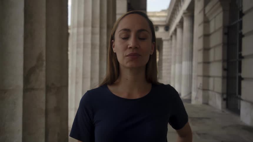 A woman stands next to a pillar staring off to the left then turns to smile at the camera