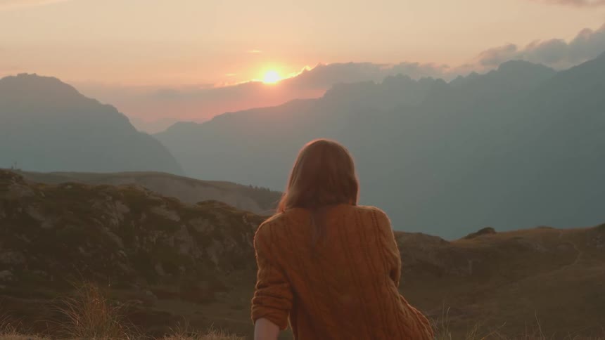 Woman sitting on the slopes of a mountain peak looking at the sunset over the clouds