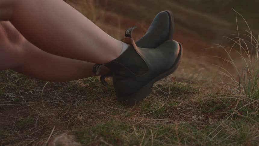A woman with bare legs and boots sitting on a grassy hill