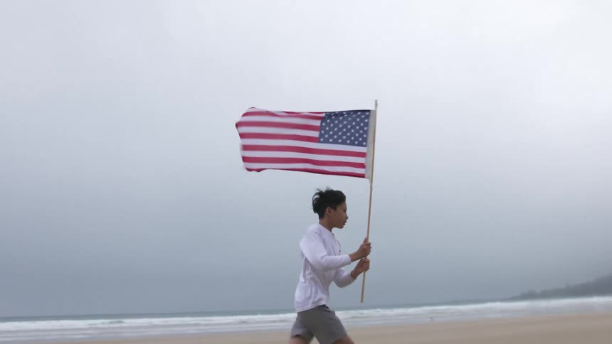 A teenage boy runs along a beach holding an american flag in foggy conditions
