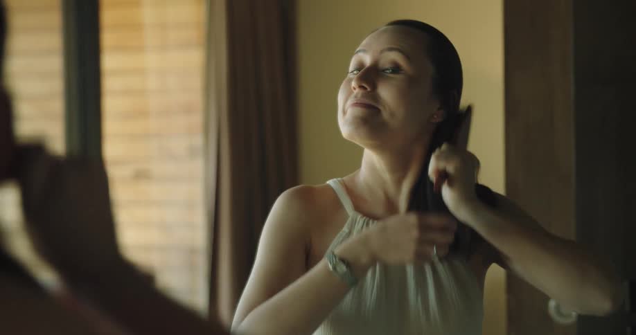 Woman brushing her long dark hair in front of a mirror in a room at home