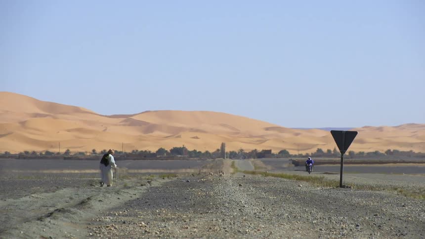 An arab man in traditional dress uses a walking stick as he walks along a road toward desert sand