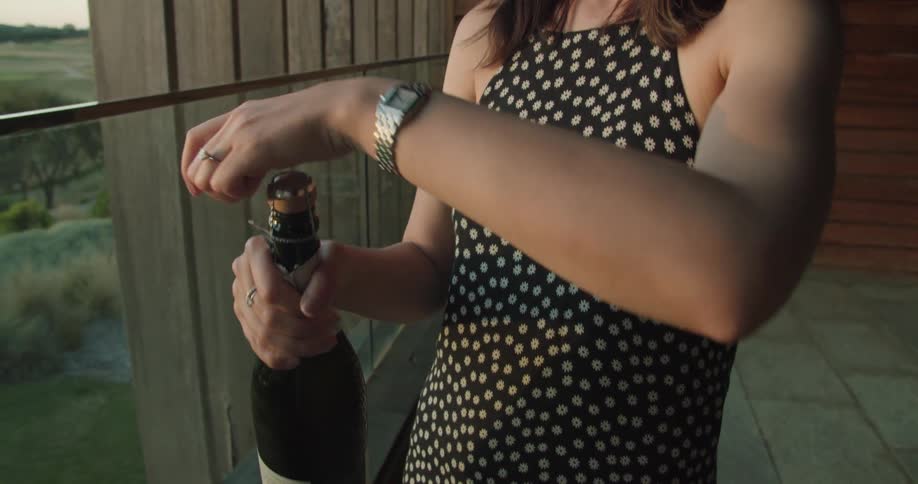 A woman in a black polka dot dress stands next to a large window at her house and removes the foil from a bottle of wine