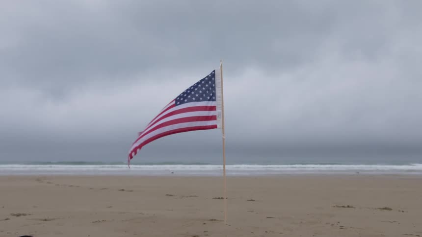 A beach with an american flag planted in the sand with the ocean sky and clouds in the background