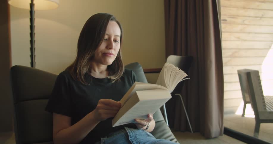 Woman leisurely reading a book sitting in a chair in the livingroom at home