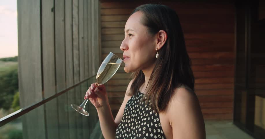 A woman stands on her balcony and smiles as she drinks a glass of champagne