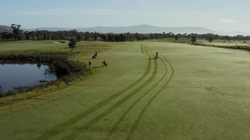 A trio of golfers walking along the fairway pause to look for their ball at the edge of a water hazard