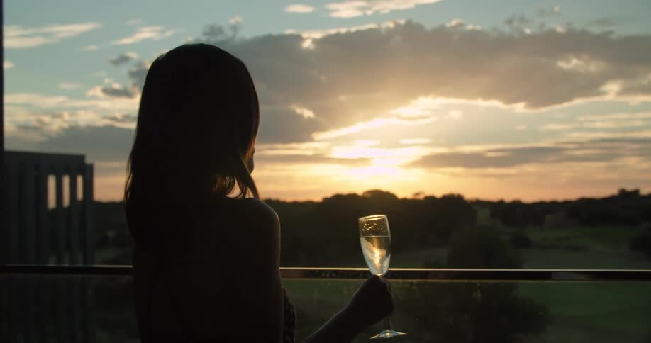 A woman stands on her balcony and drinks champagne as she looks out at the land