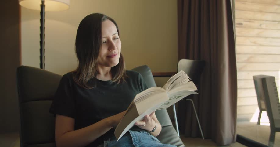 A woman sitting on a couch reading a book with a lamp and a table in the background