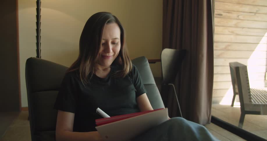 A woman smiles as she writes sitting in a chair near patio doors