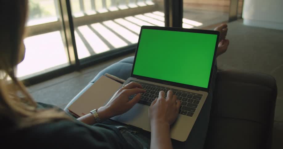 A female is sitting in a chair near a glass sliding door typing on a small laptop