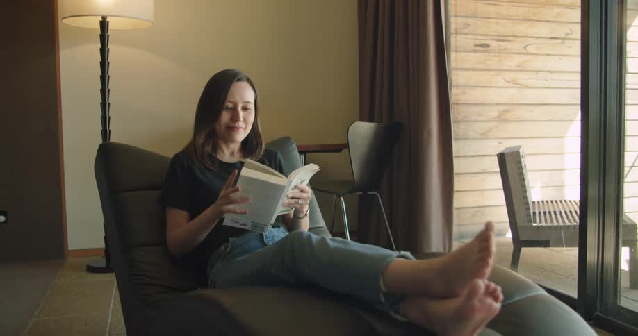 A woman smiles softly as she picks up a book to read while reclining in a chair