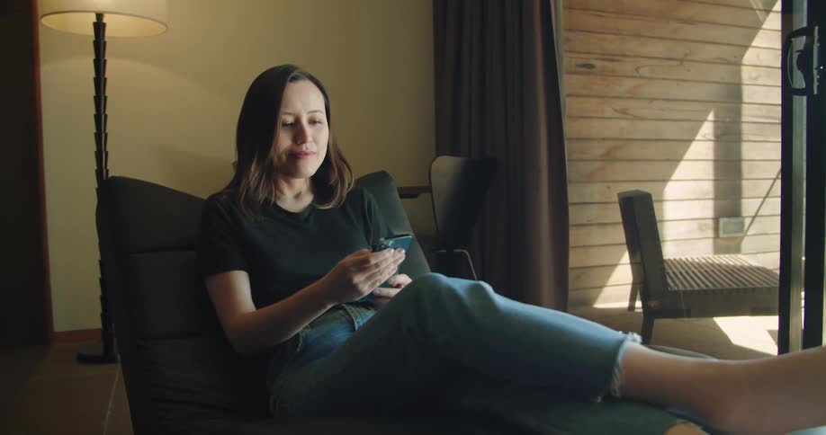 A woman sits with her legs up next to a sliding glass door indoors and takes out her smartphone