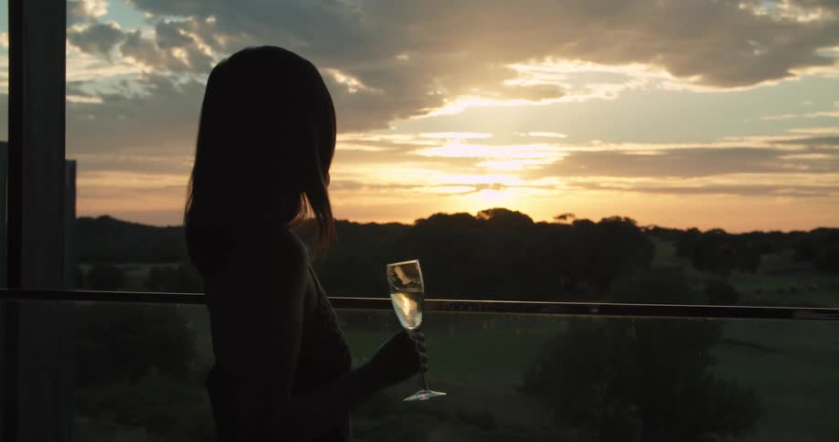 A woman drinks wine while looking at the sunset from her balcony and walks away
