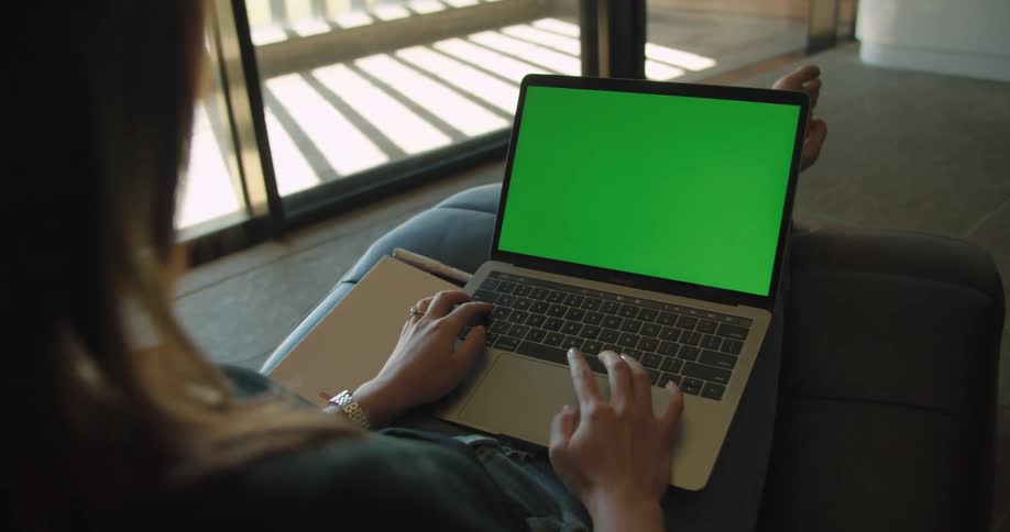 A woman sits with a laptop on her lap with a green screen indoors