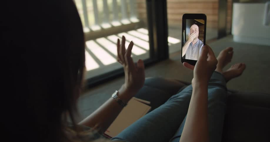 A woman sitting on a lounge chair in a house waves and blows a kiss to a man through video chat on her smartphone