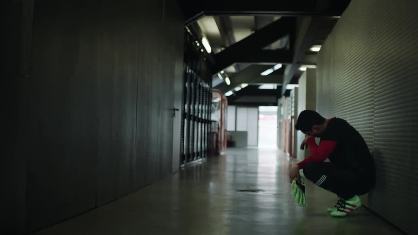 Man crouches in empty hallway of large building holding gloves and wiping under his eyes
