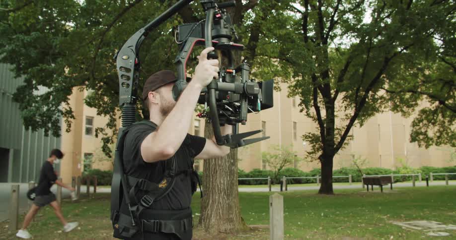 A man with a hat holding a camera and equipment with trees and a building in the background