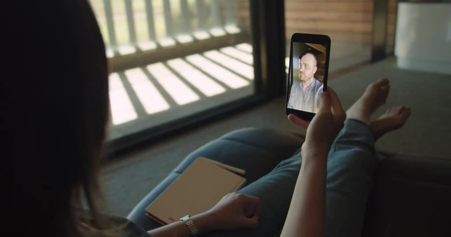 A woman sits on a chair in her apartment while talking on her smartphone with a man via video call