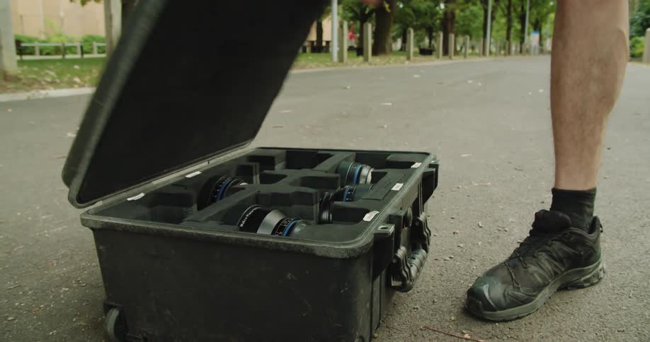 A man on a paved trail in a park removes a large case from a wheeled cart and opens it to reveal camera lens