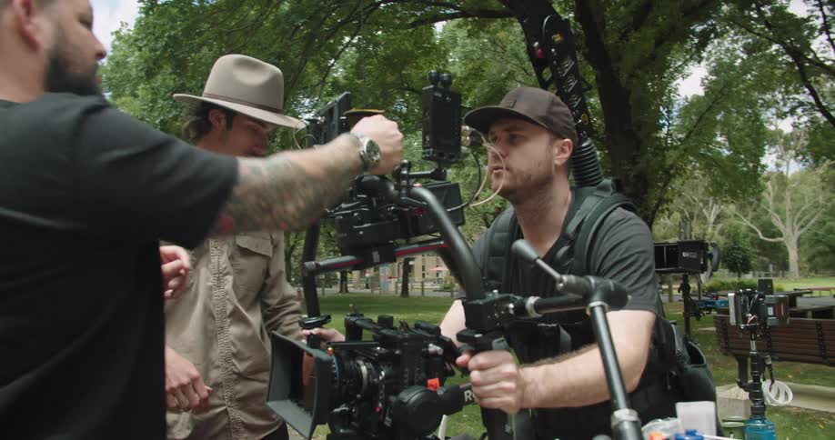 Man sits near bench watching as camera crew adjusts camera in park near buildings