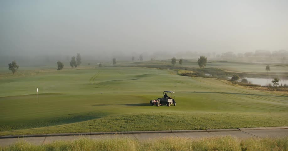 A person who maintains the golf course is slowly driving around it in a golf cart while towing equipment