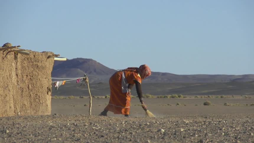A woman sweeping the ground with a hand broom in the desert with the sky and clouds in the background