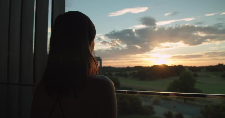 A woman stands looking out of a window at the landscape with the sun setting while holding a wine glass