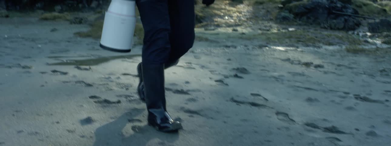 Photographer wearing rubber boots walks on the wet sand with a camera long focus lens