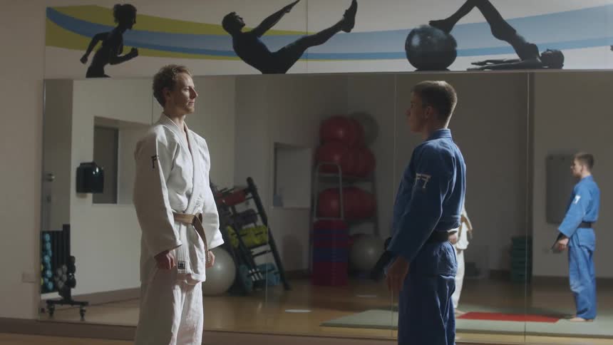 Two men wearing judo uniforms bow and start fighting on a gym in front of a mirror