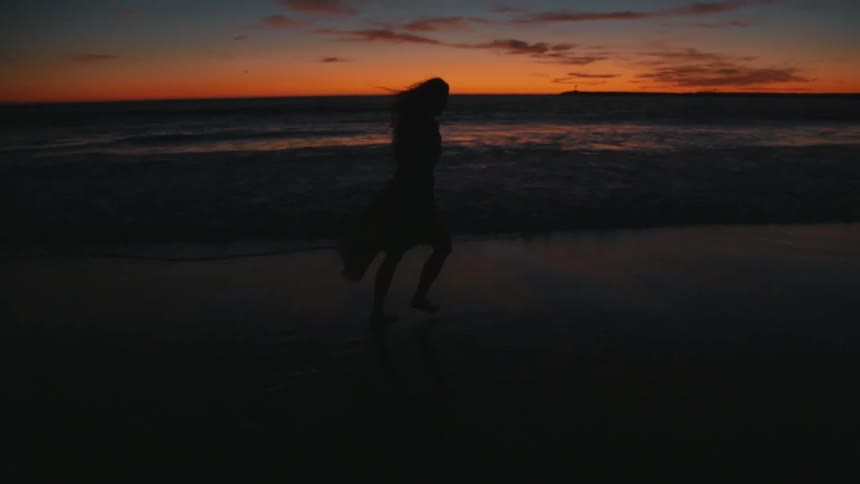 Woman runs across beach wading in and out of surf silhouetted by a red sky