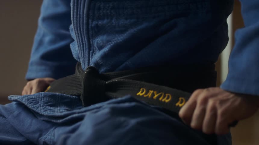 Martial artist sitting on the floor adjusts black belt over blue judo uniform