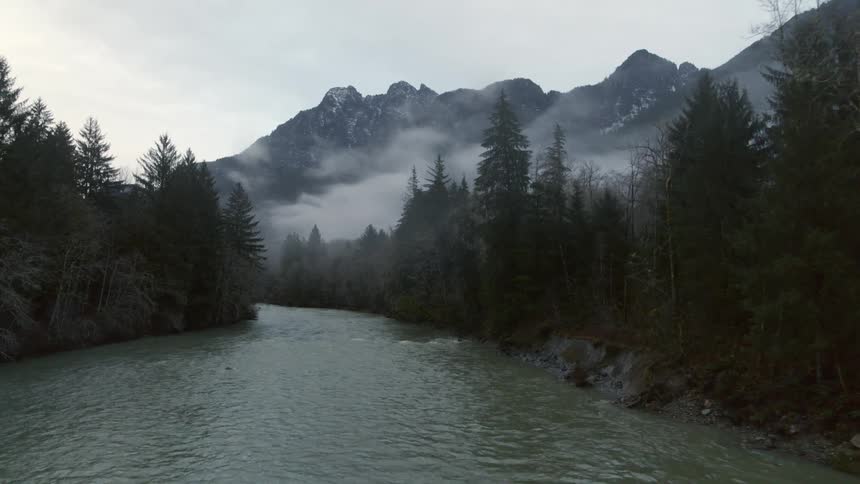 Traverse above an alpine river in the woods as fog shrouded mountains loom high in the distance