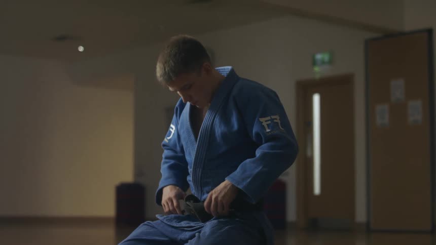 A man in a room kneeling on the floor tightening his belt wearing a martial arts gi